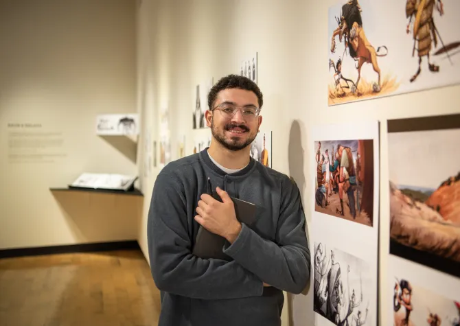 A man holding a text book leaning up against a wall with art looking confidently at the camera