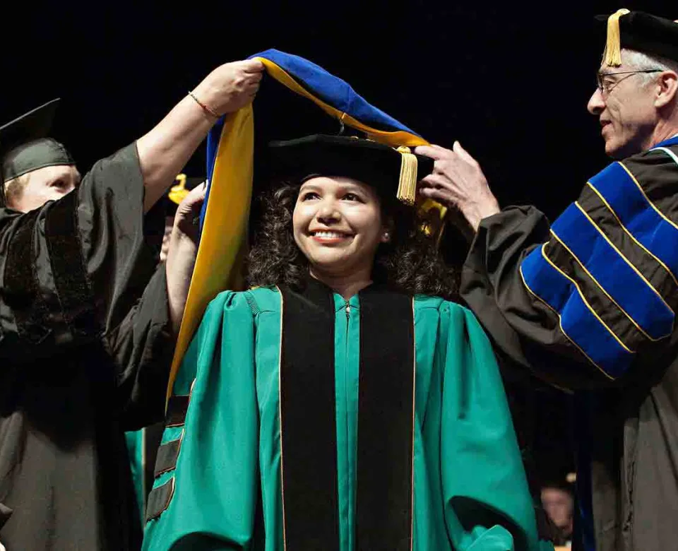 Student receiving graduate hood from Dean and advisor during commencement ceremony.