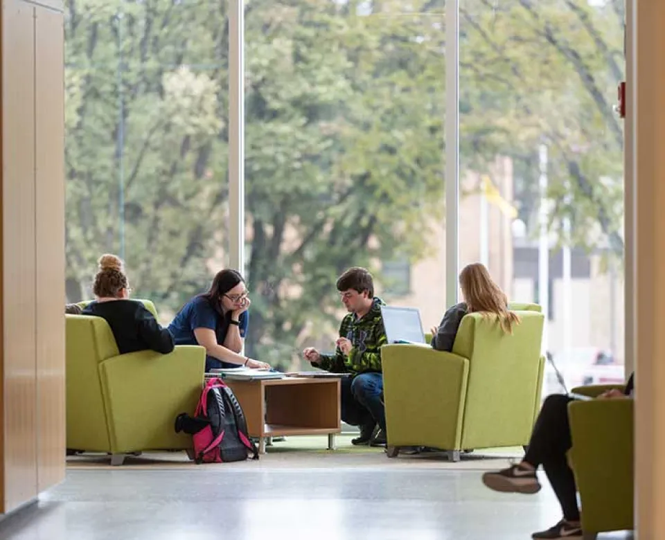 Four students studying around shared table in front of full-length windows in A. Glenn Hill Center.