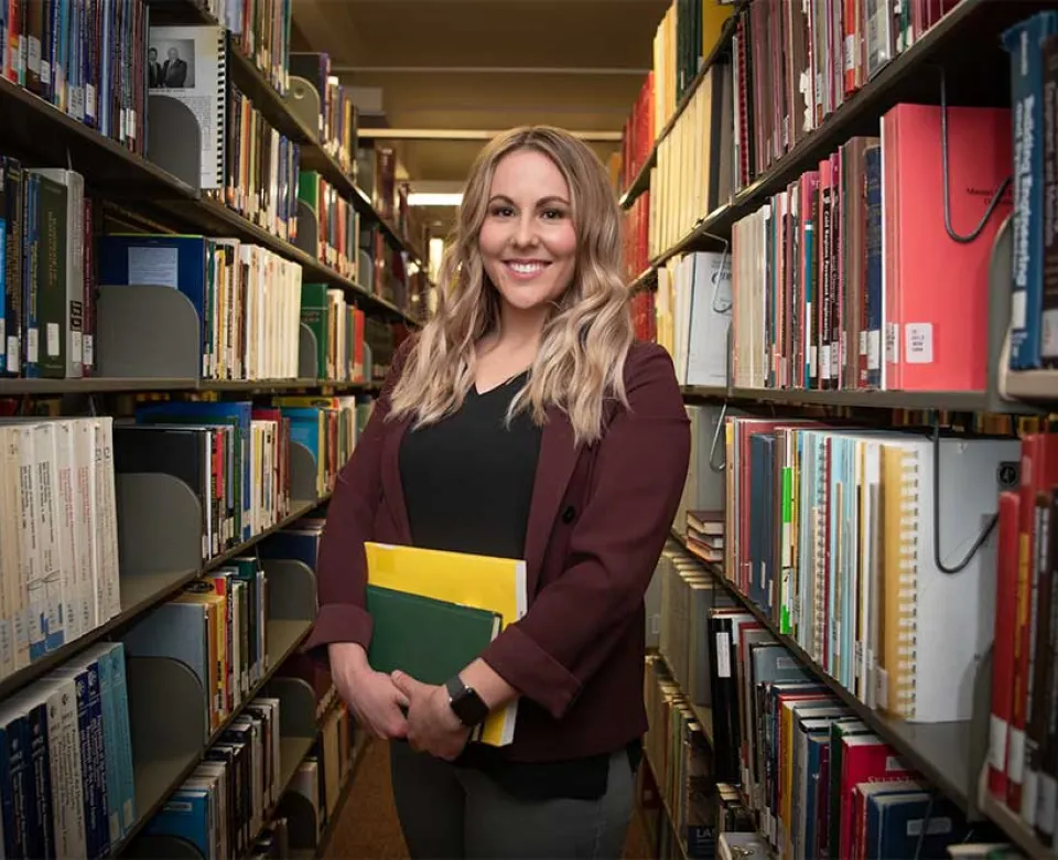 Student standing is aisle between two rows of bookshelves in the library.