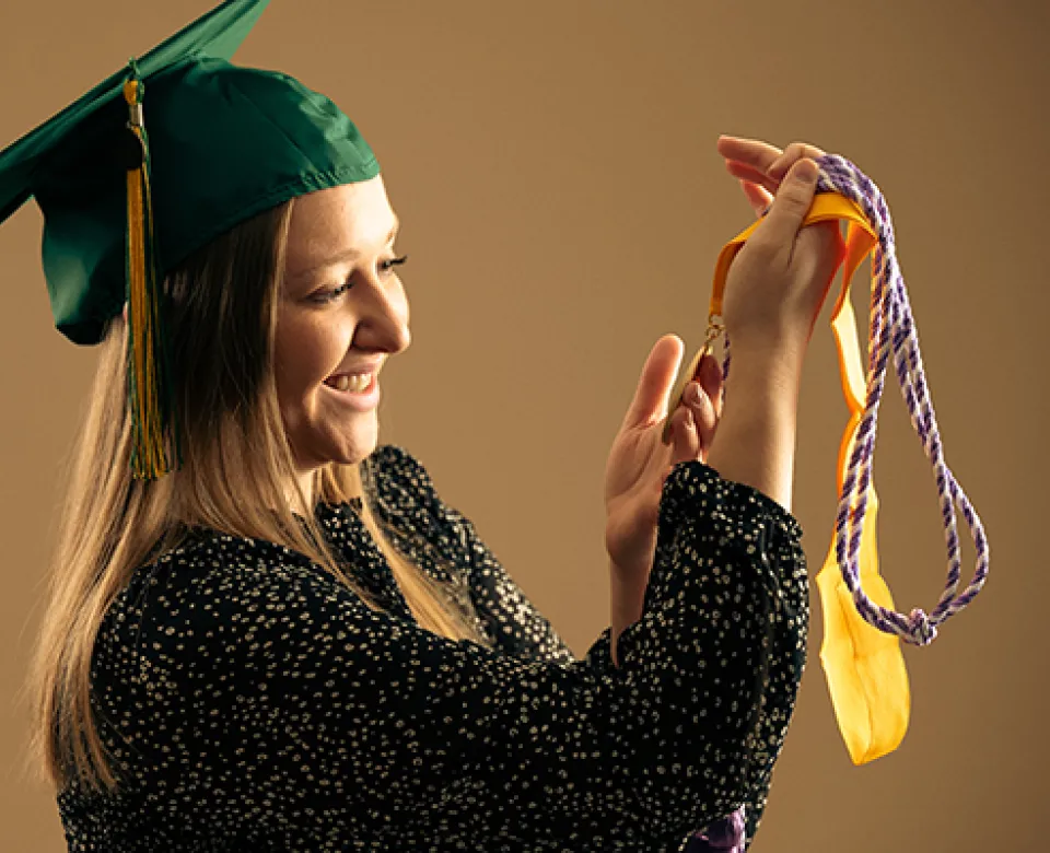 Student wearing mortarboard looking at graduation cords and medallion.