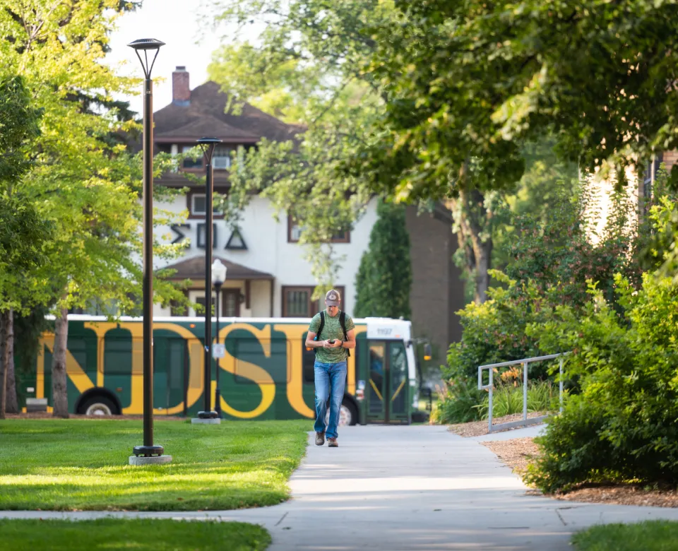Student walking down sidewalk as campus bus passes in background.