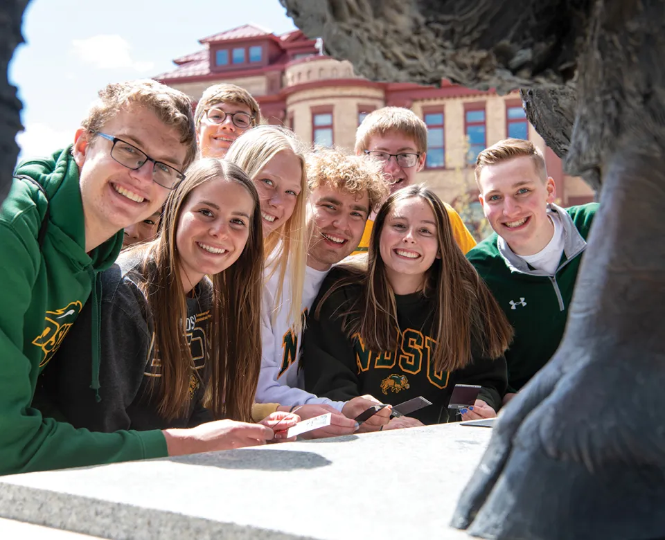 Group of eight students posing with front leg and beard of Bison statue in the foreground and Minard Hall in the background.