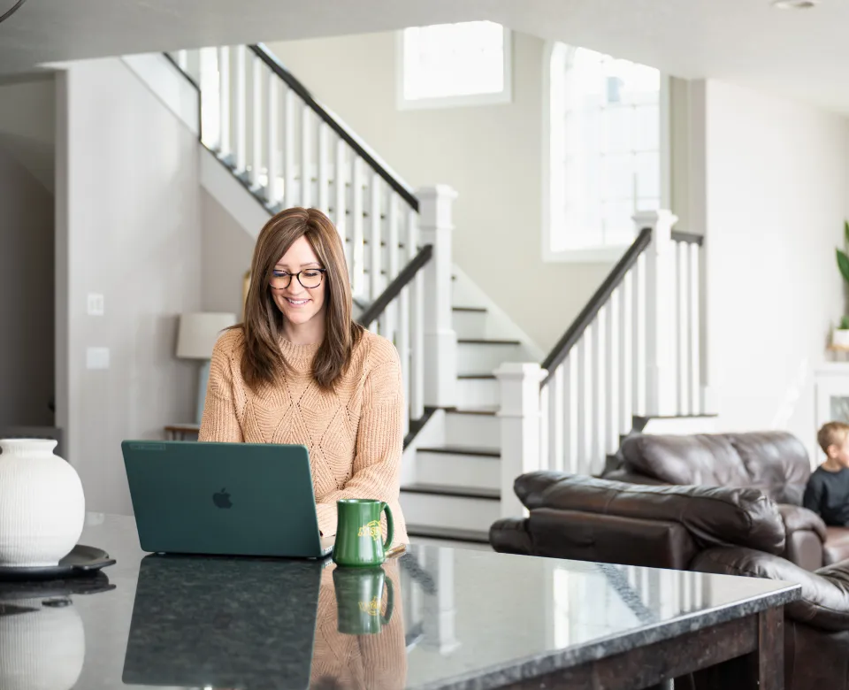 Woman sits at kitchen island in her home with a laptop computer and a green mug