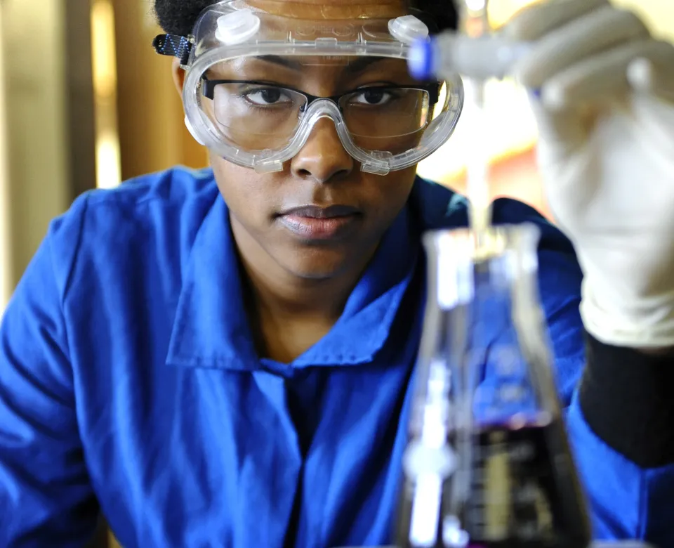 Student researcher wearing a blue shirt and lab goggles looking intently at a beaker containing a dark purple liquid