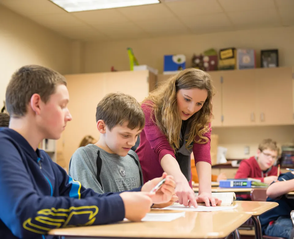Student teacher leaning on student desk explaining and pointing at students paper.