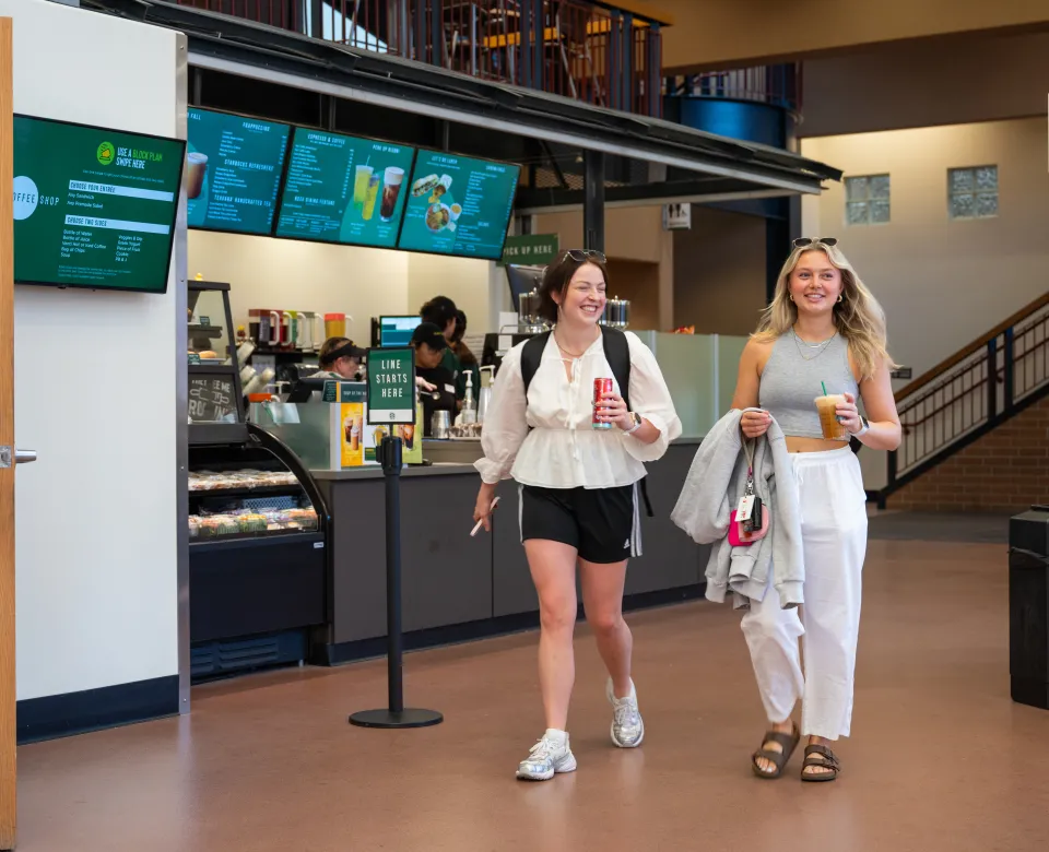 Two female students walking through a building. One is carrying a cold beverage and the second is enjoying an iced coffee.