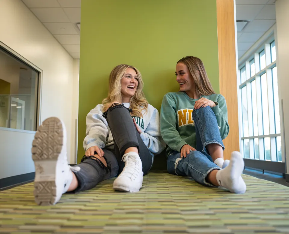 Two students sitting up against a green wall talking and smiling.