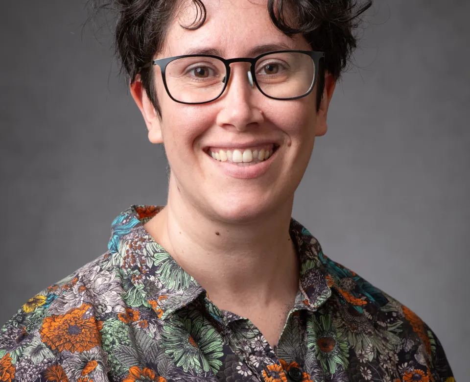 Evelyn Milburn, a white woman with brown hair and glasses is wearing a bright collared shirt and smiling broadly.