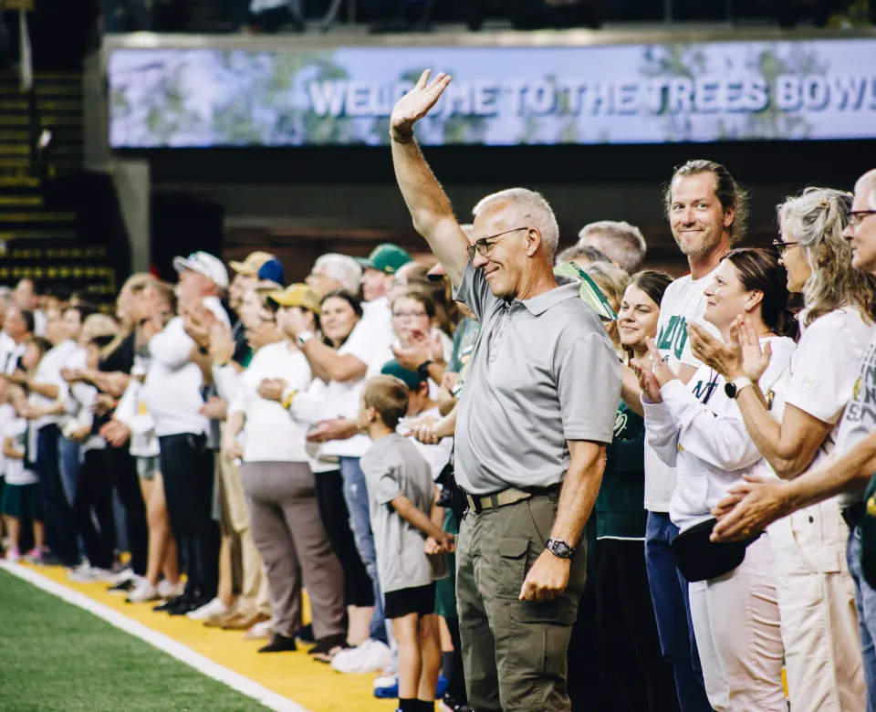 A man raises his hand to wave on field at a football game next to a group of people in line