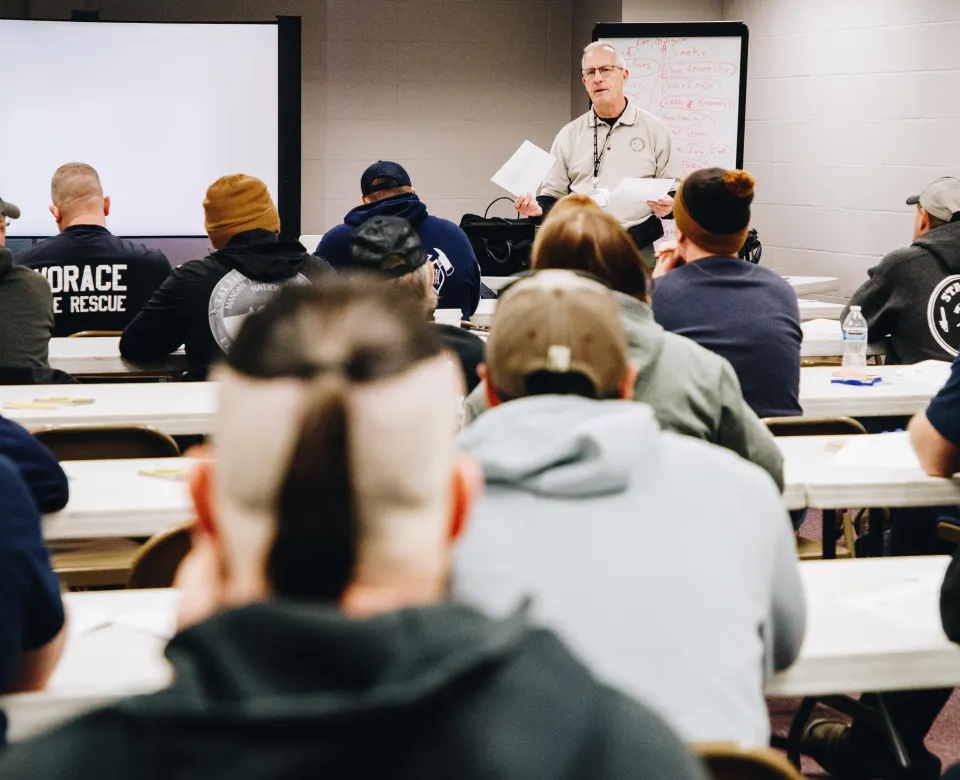 A man leads a class for many firefighters sitting at tables