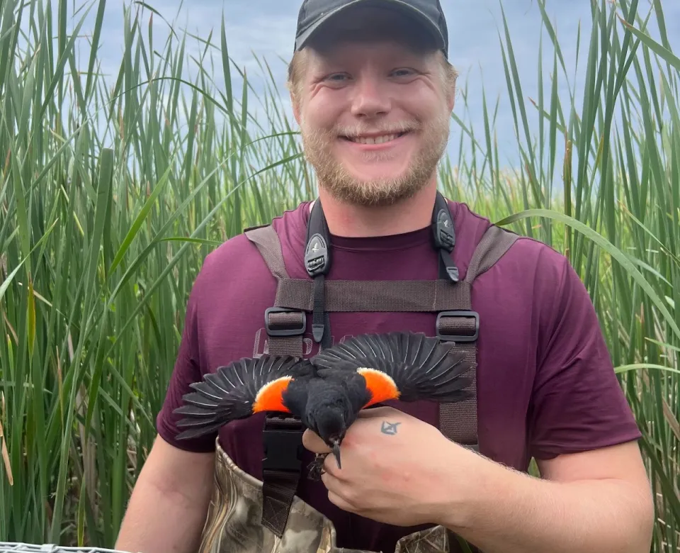 Koby holding a bird, standing in a grassy field, smiling confidently at camera. 