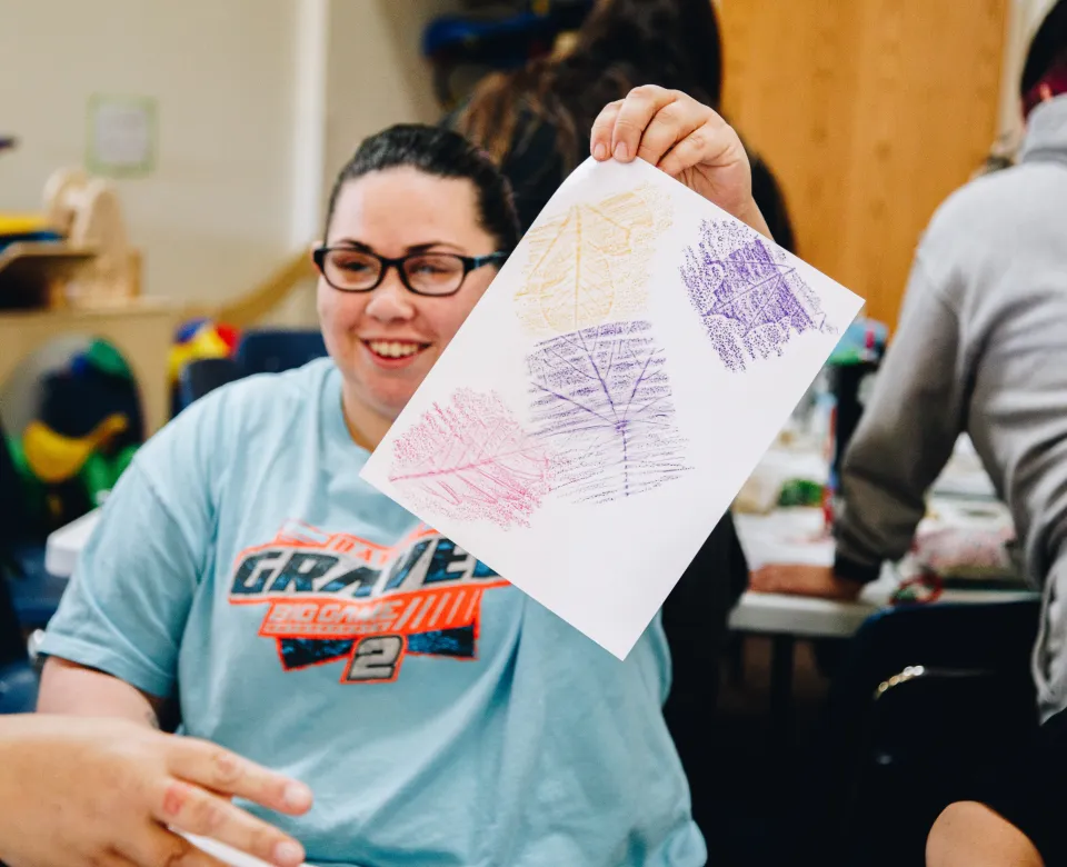A woman in glasses holds up a piece of paper with leaf patterns made using crayons on it