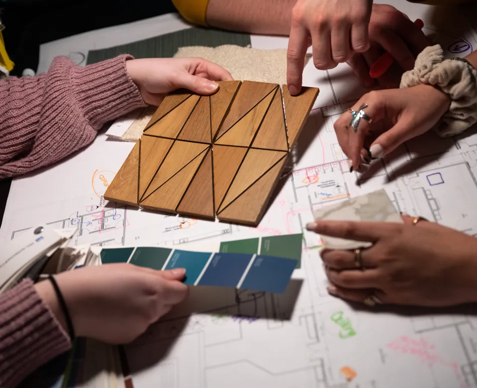 Three sets of hands working with color samples and tile samples on top of a floor plan rendering.