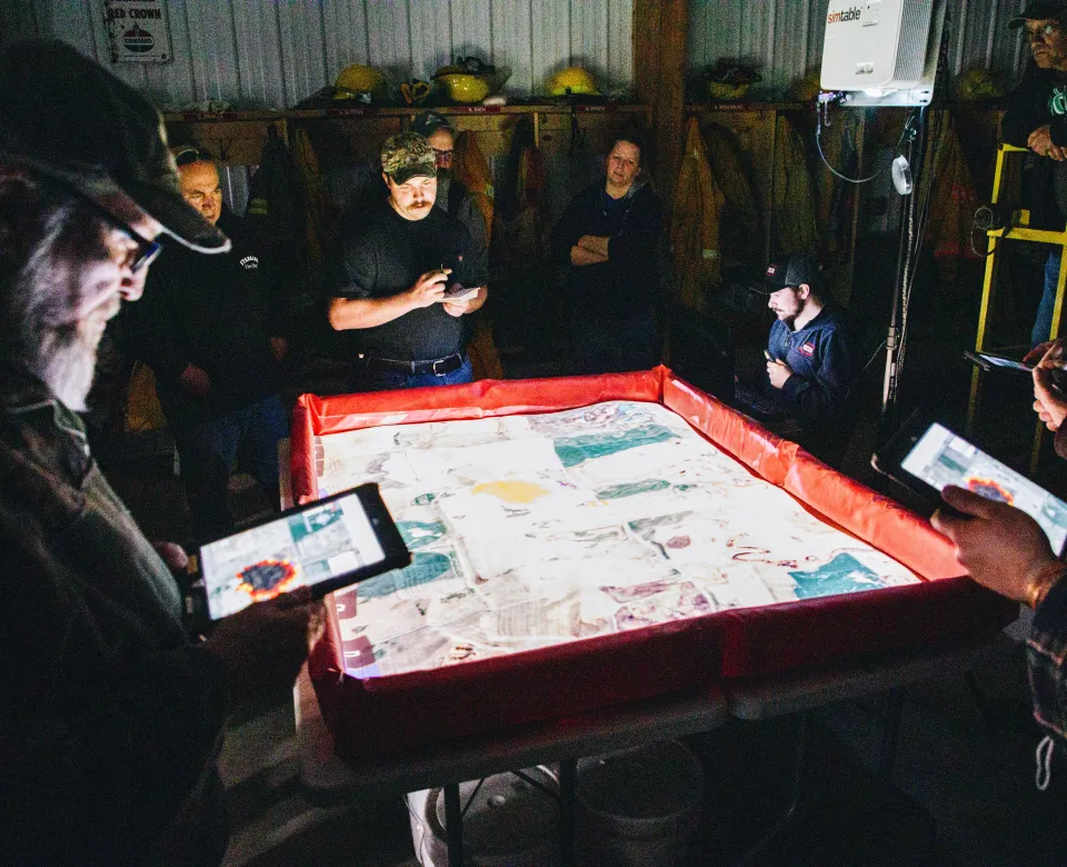 A group of volunteer firefighters use a virtual simulation table to practice incident command systems during a simulated wildfire scenario