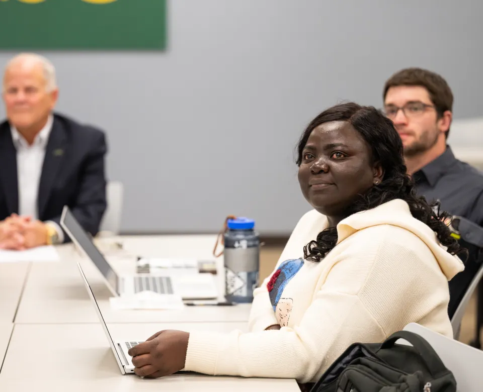 Students sitting at table with laptops open while listening to someone speak.