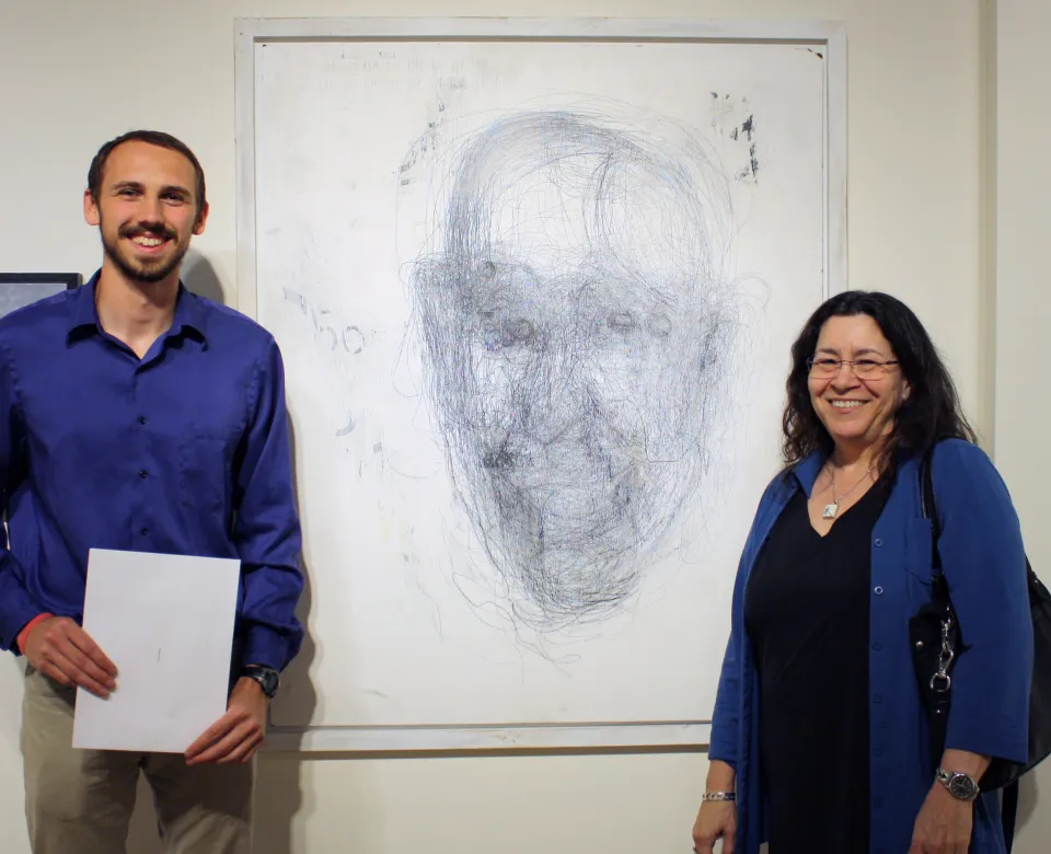 A young man and woman stand in front of a pencil drawing of a face. 