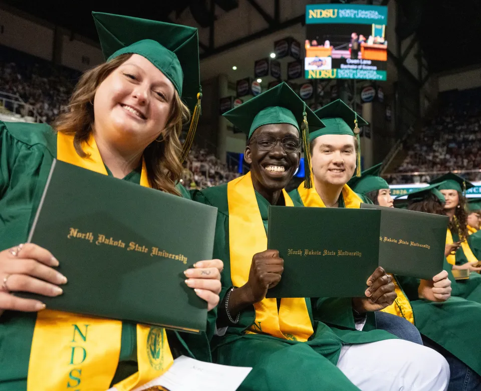 NDSU graduates wearing cap and gowns hold up their diplomas