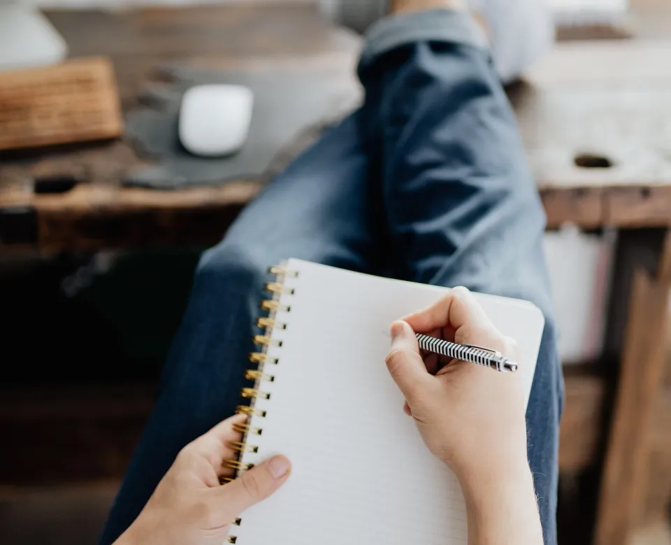 Person with feet up on a desk, blank page of a notebook and pen in hand