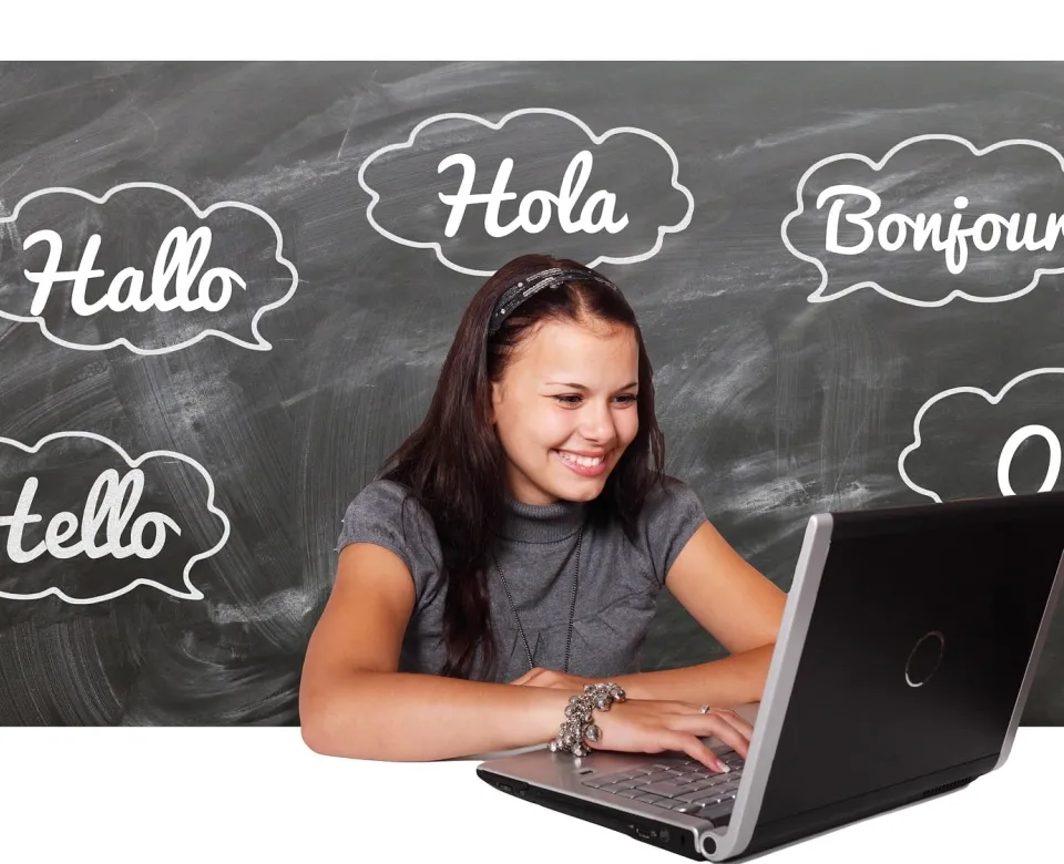 Person on laptop with a blackboard behind her and five different languages of the word Hello. 