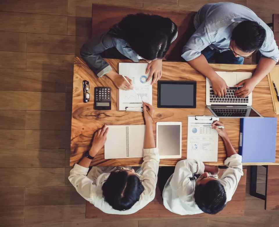 Professionals working at a table together, some working with pen and paper and one is working on laptop.