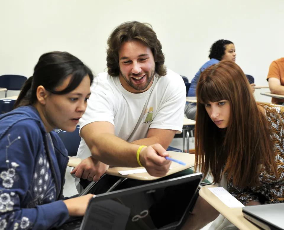 Three students in a classroom looking at information on a computer. One student is pointing and talking. 