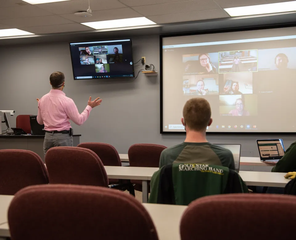 Three people in a classroom setting speaking to a group participating via Zoom.