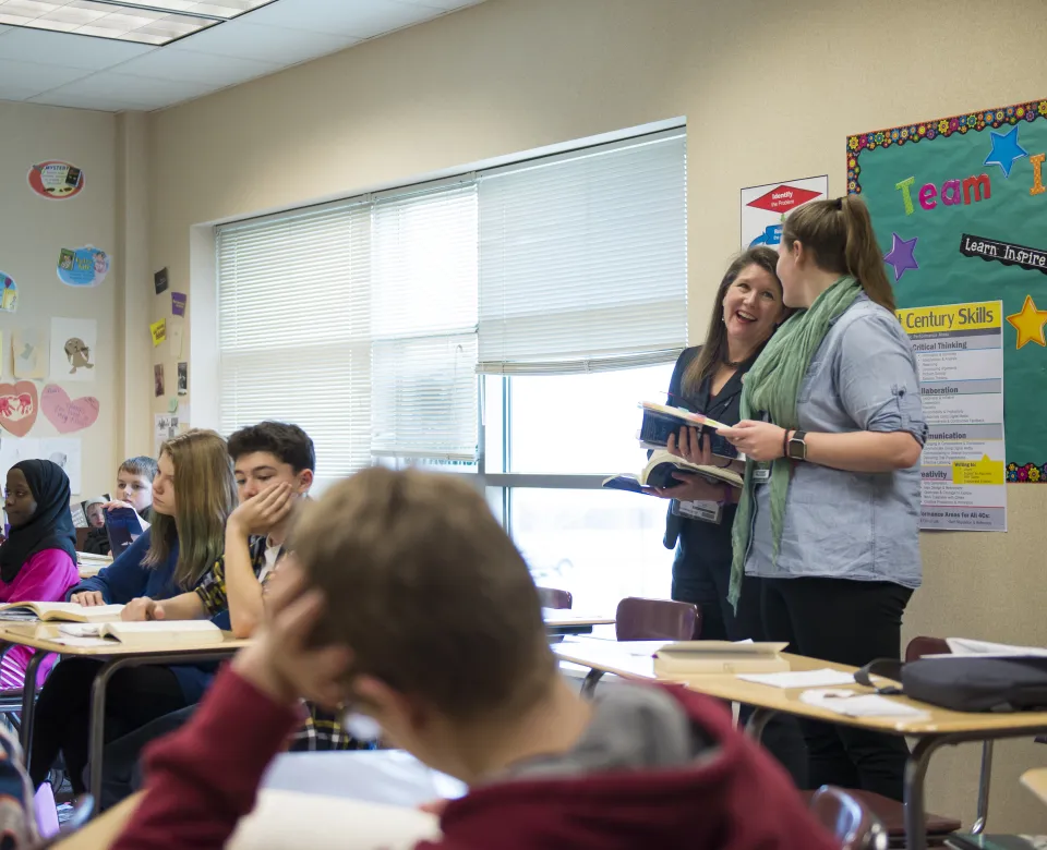 Teacher talking with a student teacher in a classroom setting with students sitting at individual desks