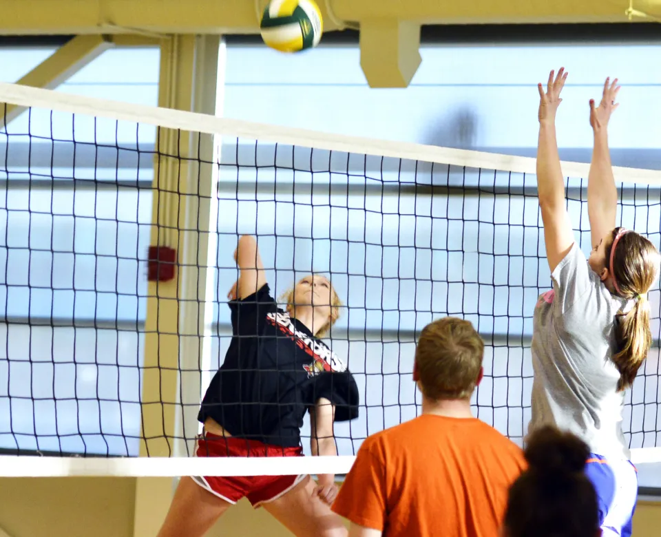 Students playing volleyball. One student is mid-air to spike the ball while another student is mid-air trying to block the spike. Other students are looking on.