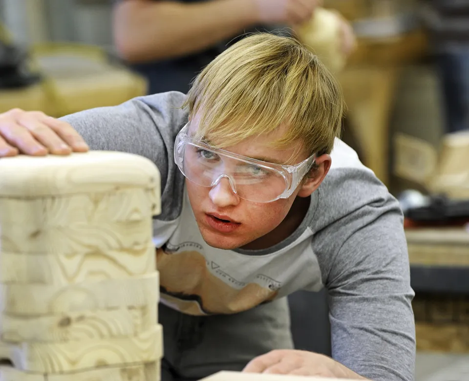 Student wearing protective eyeware while working on a wooden structure in woodshop.