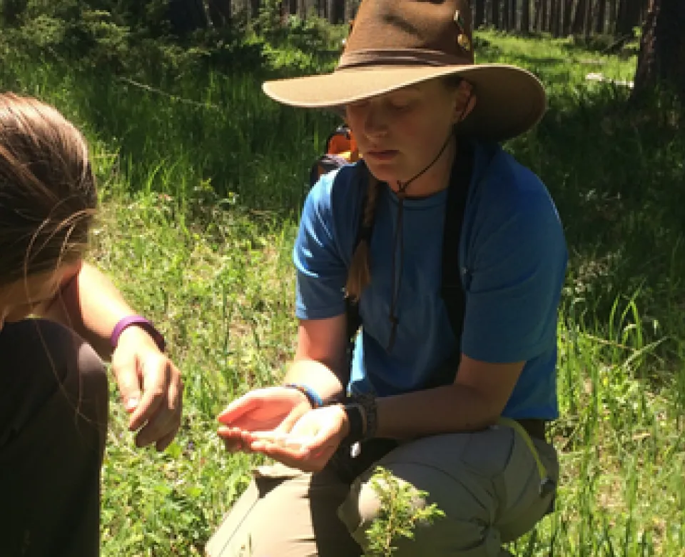 Student wearing a hat and blue shirt while working in the field. He is holding something in his hands while crouched down in the grass.