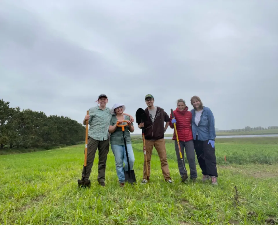 Students standing in field holding shovels and smiling confidently at camera.