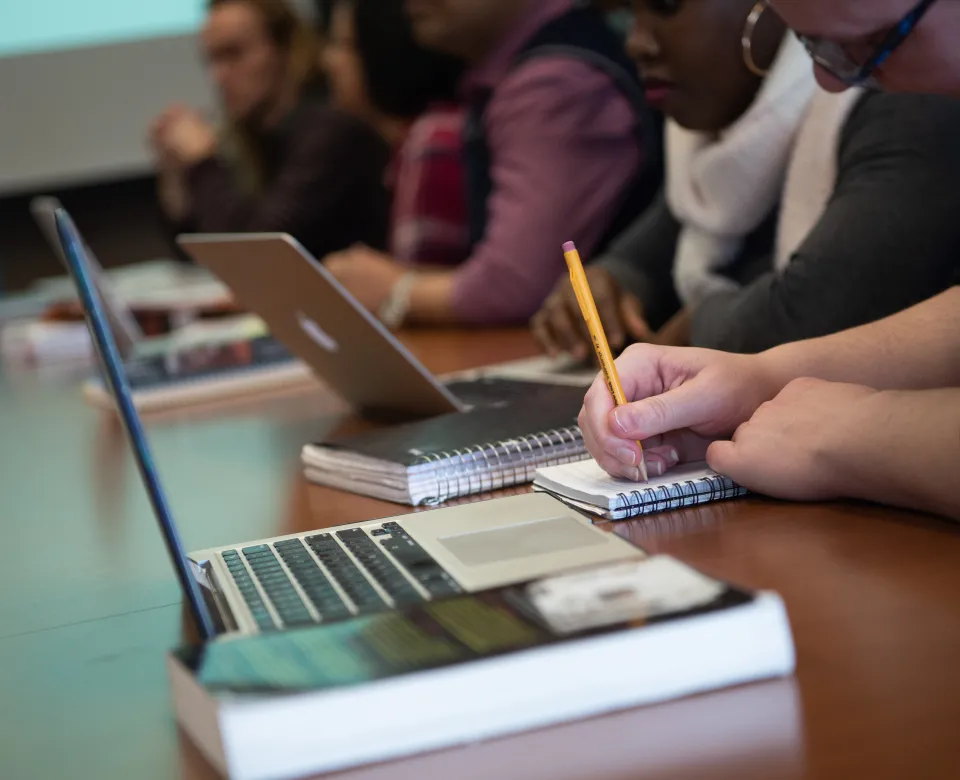 Students working with pencil and paper while sitting at table with laptops and books.