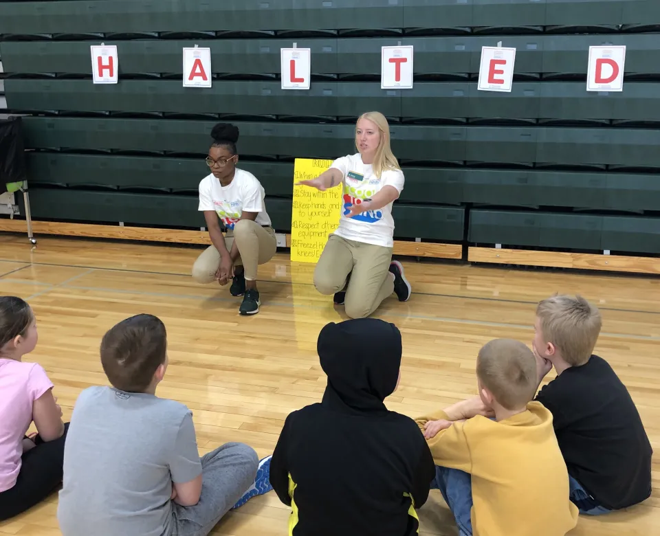 Two student-teachers speaking to a small group of students sitting on the gym floor.