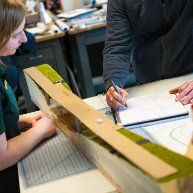 Students watching professor as he instructs using pen and paper in design studio.