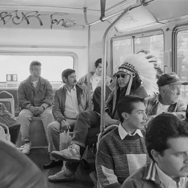 A man with a native american headdress sits on the bus among other men traveling. This image links to the artists website.