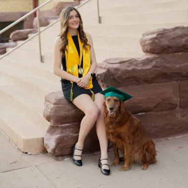 campus shot of graduate and her dog who is wearing her graduation cap