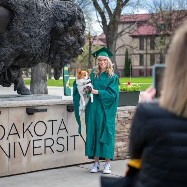 graduate holding her small dog in front of the bison statue