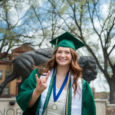 student posing in front of the Bison Statue wearing her graduation cap and gown