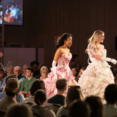Two models wearing floor-length prom dresses and white gloves.