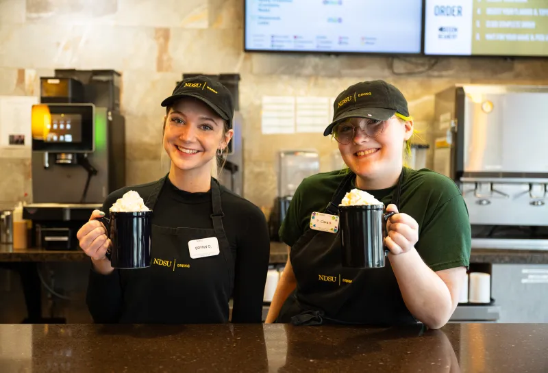 Two female dining students working the bison beanery posing with two black coffee mugs topped with whipped cream