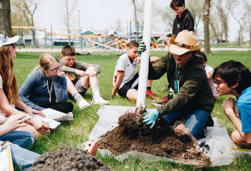 A woman in a cowboy hat shows a group of middle school students how to plant a tree
