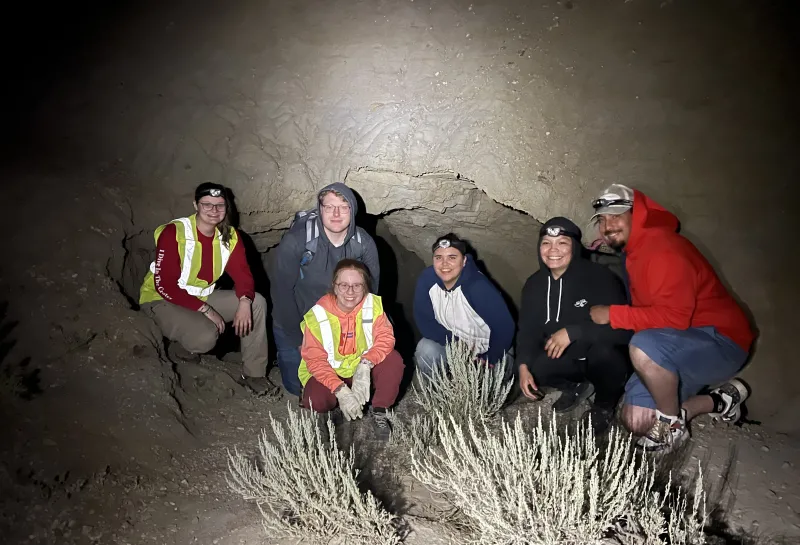 Students stitting in front of rock/cave smiling confidently at camera. 