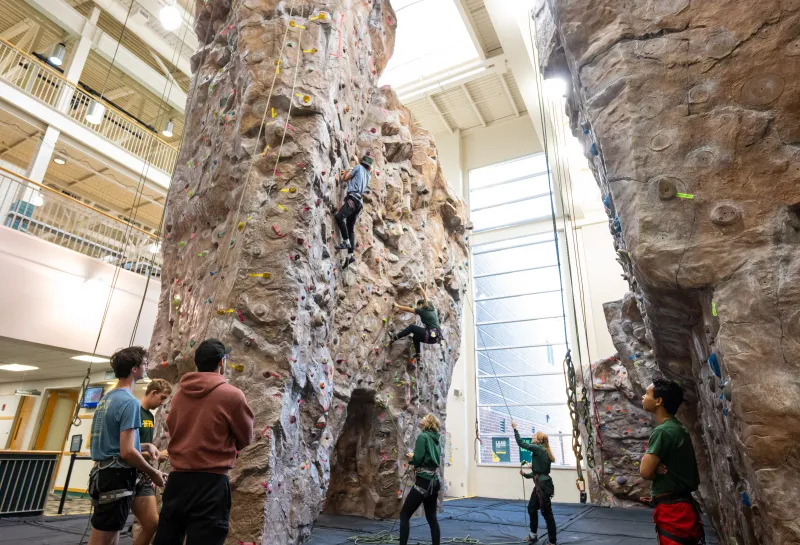Students watching climbers on rock wall and others belaying climbers