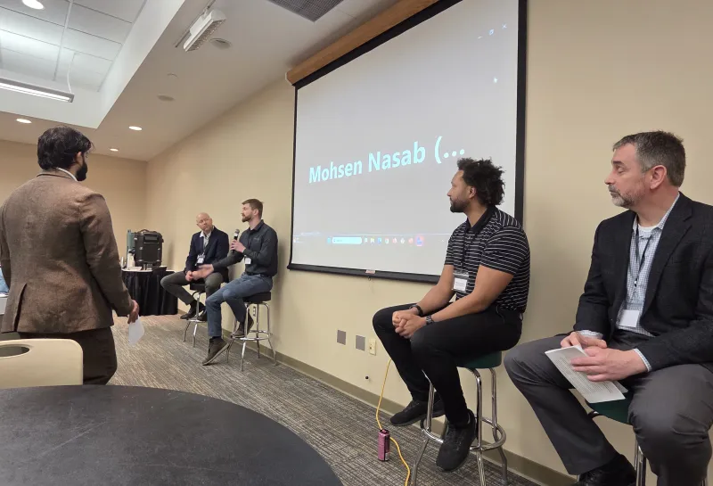 A panel discussion in a conference room with four men, three seated on stools near a projection screen 