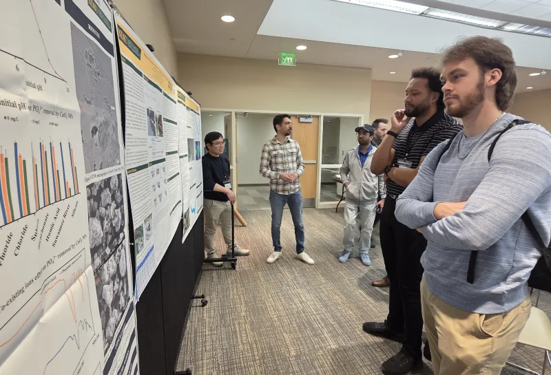 Academic poster session showing several people standing and observing scientific posters displayed on black boards.