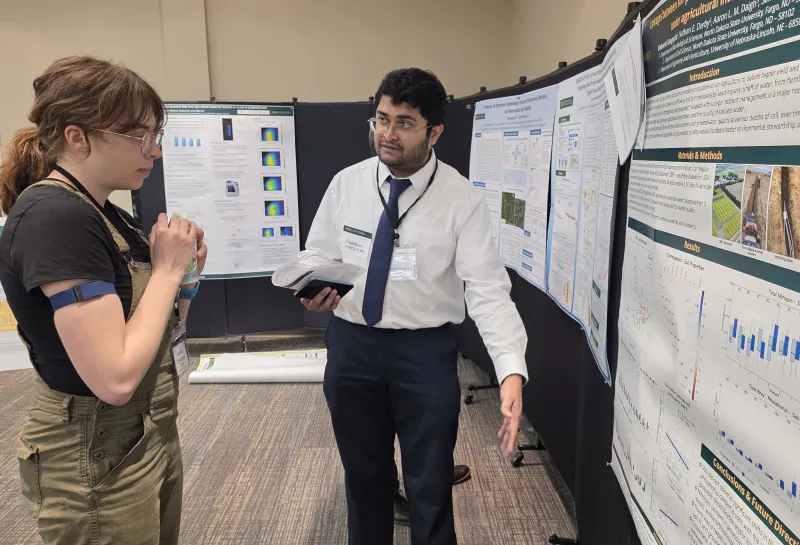 Two people engaged in discussion at a scientific poster presentation, with multiple posters displaying charts, graphs, and images related to research displayed around them.