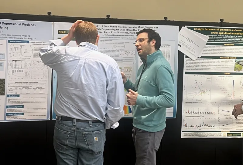 Two people discuss scientific research posters displayed on a black partition wall at a conference or academic event. The posters feature detailed charts, graphs, and text on topics including wetland processes, machine learning models, and soil properties.