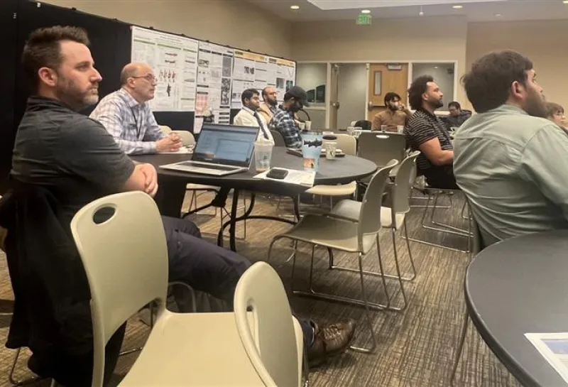 A seminar showing attendees seated and facing forward, likely listening to a presentation. Posters with charts and graphs are displayed on the back wall, and several attendees have laptops open, indicating a professional or academic setting focused on data or research.