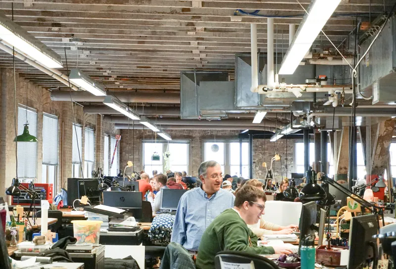 Architecture studio with students working at large tables with computers and other supplies with an instructor looking on.